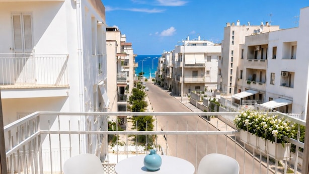 A balcony with two white chairs and a table, overlooking a street with buildings and a clear blue sky. - Dimora Peppino, a family friendly holiday home, just few step from the sea. (Polignano a Mare)