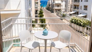 Un balcon avec deux chaises blanches et une table, donnant sur une rue bordée d’immeubles et sous un ciel bleu dégagé.