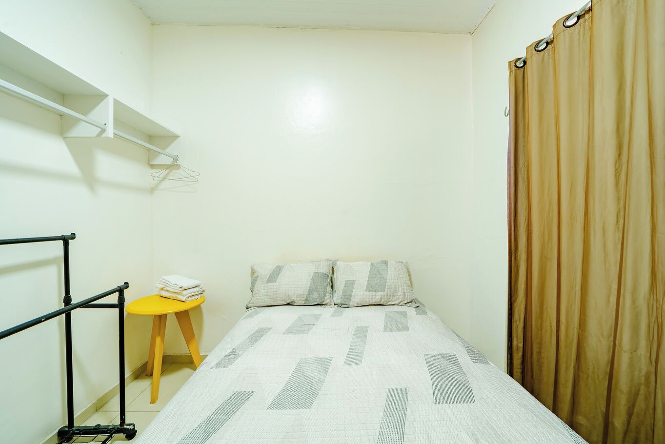 A small, single-bed room with a bed, a small table with a chair, a shelf, and a curtain.