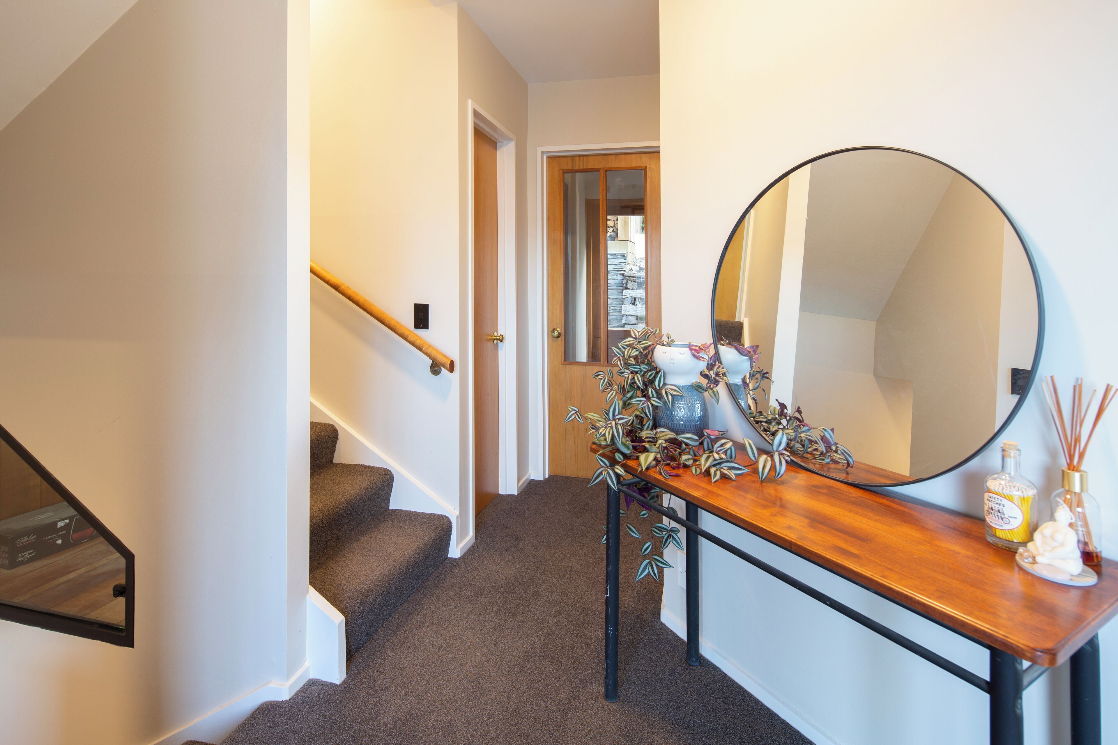 A staircase with a wooden handrail, a wooden console table with a mirror, and a vase with decorative plants.