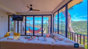 A spacious living room with a large sofa, a yellow armchair, and a ceiling fan. A large window offers a view of a mountainous landscape. - PISCIS CONDO
Dawn overlooking the most beautiful bay in Mexico. (Zihuatanejo)