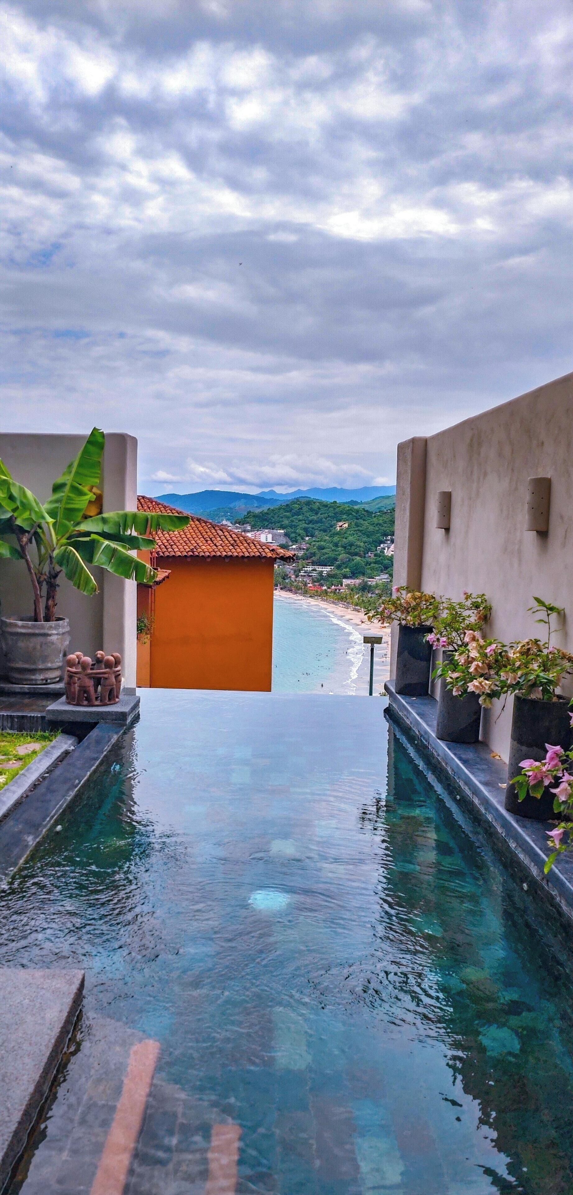 A pool area with clear water, surrounded by potted plants and a view of a beach and mountains in the distance.