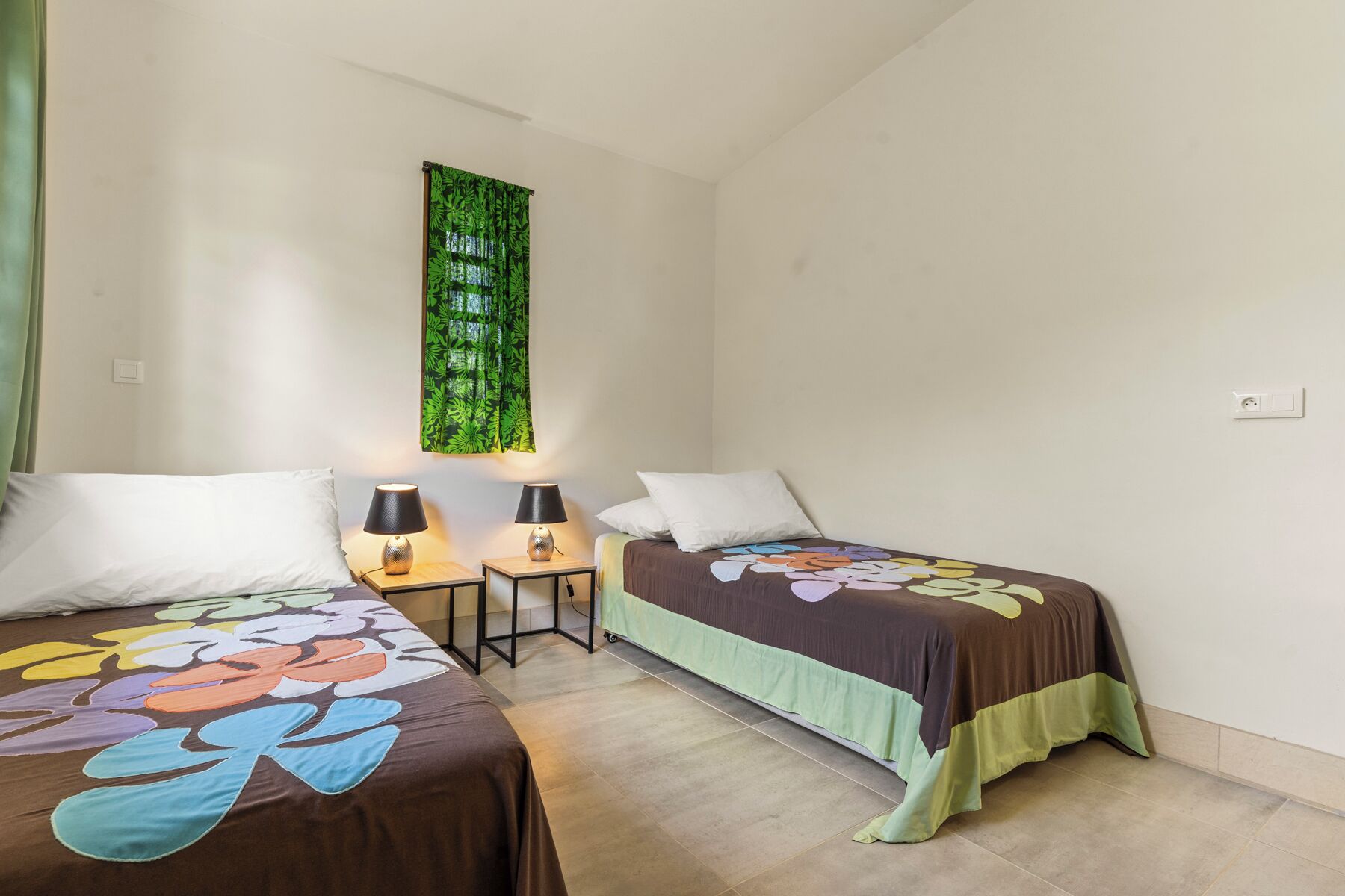Un dormitorio con dos camas, cada una con una colcha estampada con motivos florales, dos lámparas de noche, un tapiz de hojas verdes en la pared y una mesita de noche pequeña.