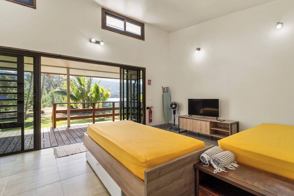 A modern bedroom with two beds, a TV, and a balcony with a view of greenery.