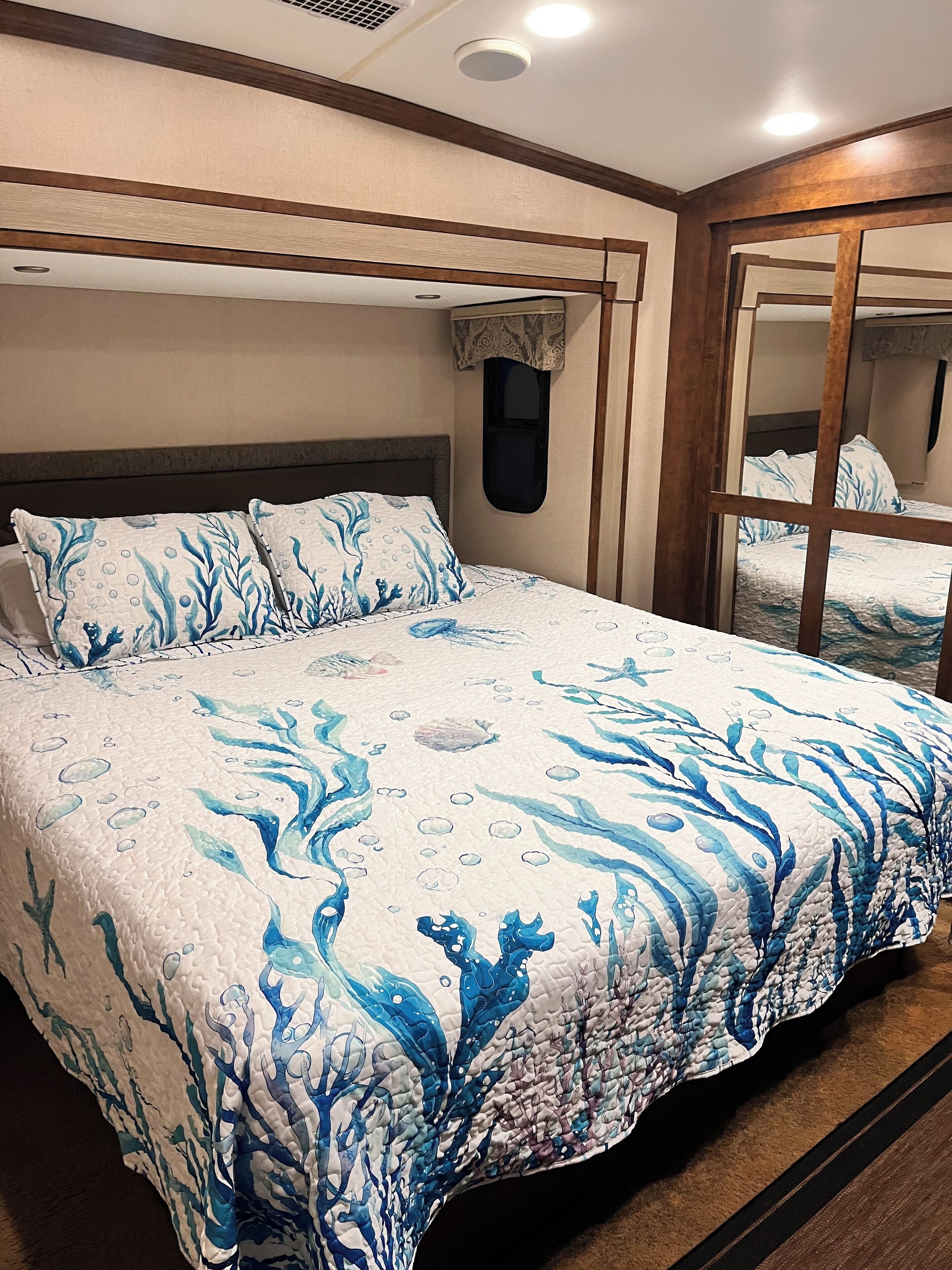 A bed with a blue and white ocean-themed bedspread, a wooden headboard, and a window with a view of the room.