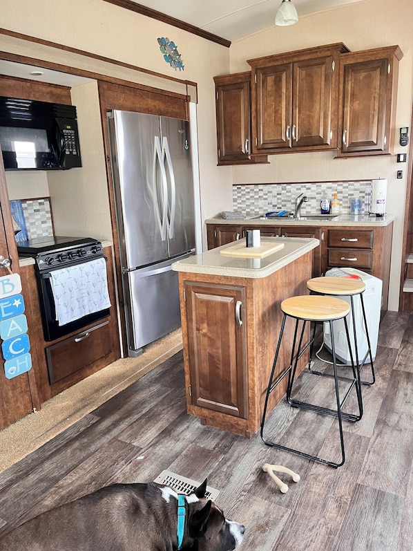 A kitchen with wooden cabinets, a stainless steel refrigerator, a stove, and a sink. There are two stools and a dog lying on the floor.