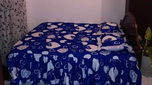 Una cama pequeña y bien hecha con colcha azul y blanca estampada, cabecera de madera y una planta en maceta a la derecha.