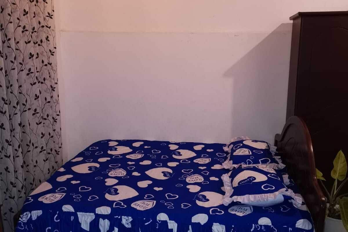 A small, neatly made bed with a blue and white patterned bedspread, a wooden headboard, and a potted plant to the right.