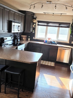 A kitchen with dark wood cabinets, a stainless steel refrigerator, a microwave, and a large island with bar stools. - ○Small Town Pearl○ 2Level Hidden Gem Near RedDeer│1K+2Q│Private Yard│Firepit+BBQ (Penhold)