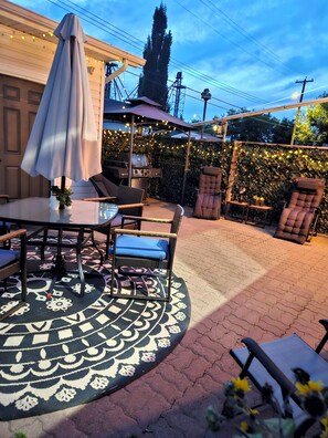 Un patio exterior con mesa, sillas, una parrilla y luces colgantes.