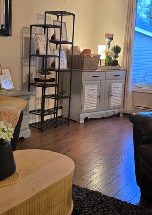 A living room with a wooden floor, a black metal shelf with framed pictures, a white cabinet with drawers, and a black leather chair.