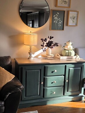 Una habitación con un armario verde, un espejo, una lámpara y objetos decorativos, incluyendo un jarrón con flores y calabazas.