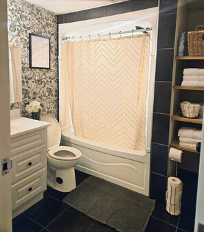 A bathroom with a toilet, a bathtub, a shower curtain, a sink, a mirror, a framed picture on the wall, and a shelf with towels and a basket. - ○Small Town Pearl○ 2Level Hidden Gem Near RedDeer│1K+2Q│Private Yard│Firepit+BBQ (Penhold)