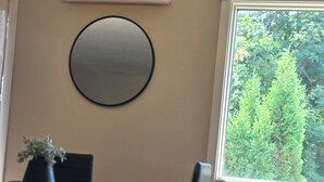A dining area with a table, chairs, a potted plant, a round mirror, and a window with a view of greenery.