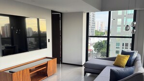 A modern living room with a large flat-screen TV, a grey sofa with yellow cushions, a wooden entertainment unit, and a view of city buildings through large windows.