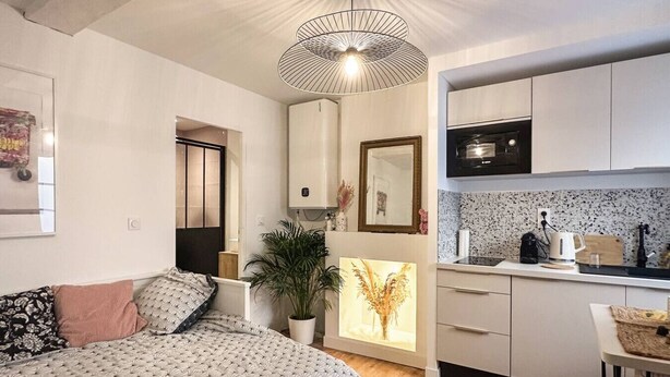 Un dormitorio compacto con cama, mesita de noche y una planta en maceta. Junto, una kitchenette con microondas, hervidor y encimera.