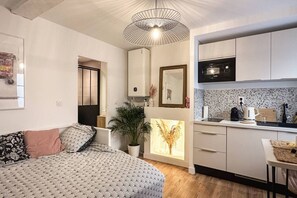 Un dormitorio compacto con cama, mesita de noche y una planta en maceta. Junto, una kitchenette con microondas, hervidor y encimera.