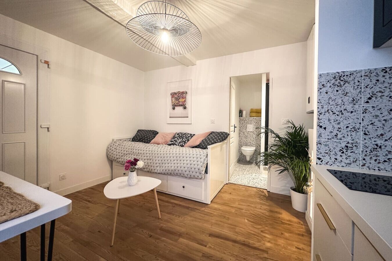 Et lille, godt oplyst værelse med en seng, et lille bord med en vase, en hængende lampe, en dør til et badeværelse og en potteplante.