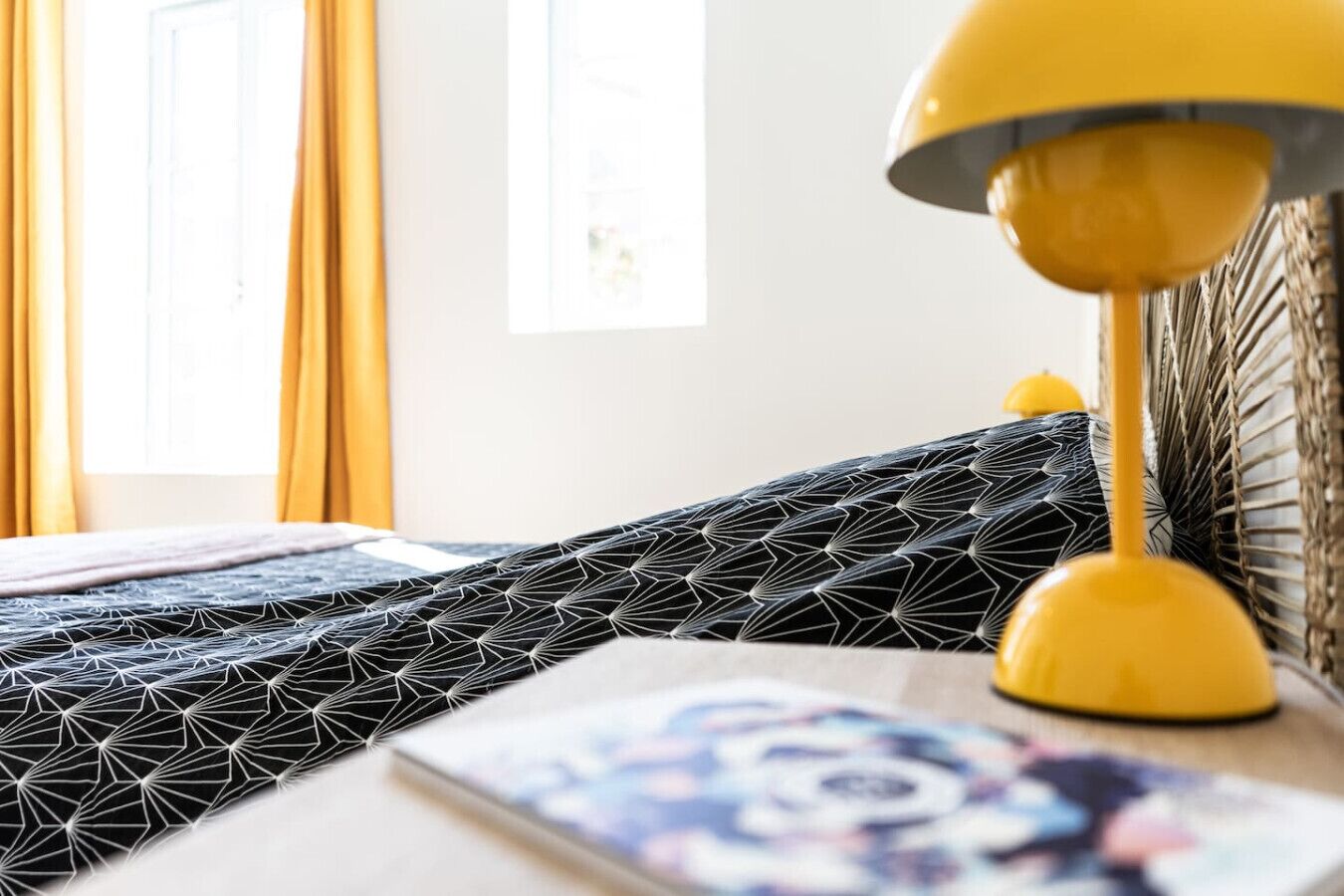 Un lit avec une couvre-lit à motifs géométriques, une lampe de sol jaune et un magazine sur une table de chevet.