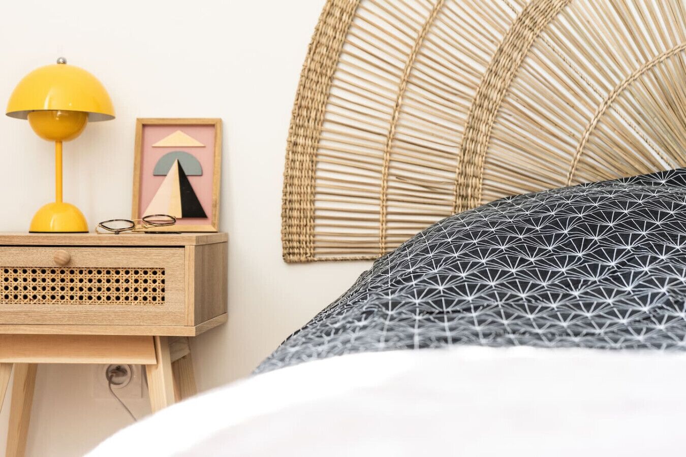 Una cama con un cojín de estampado geométrico, una lámpara amarilla y un cuadro en una mesita de noche de madera.
