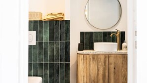 Un baño con un lavamanos blanco, un espejo redondo y baldosas verde oscuro.