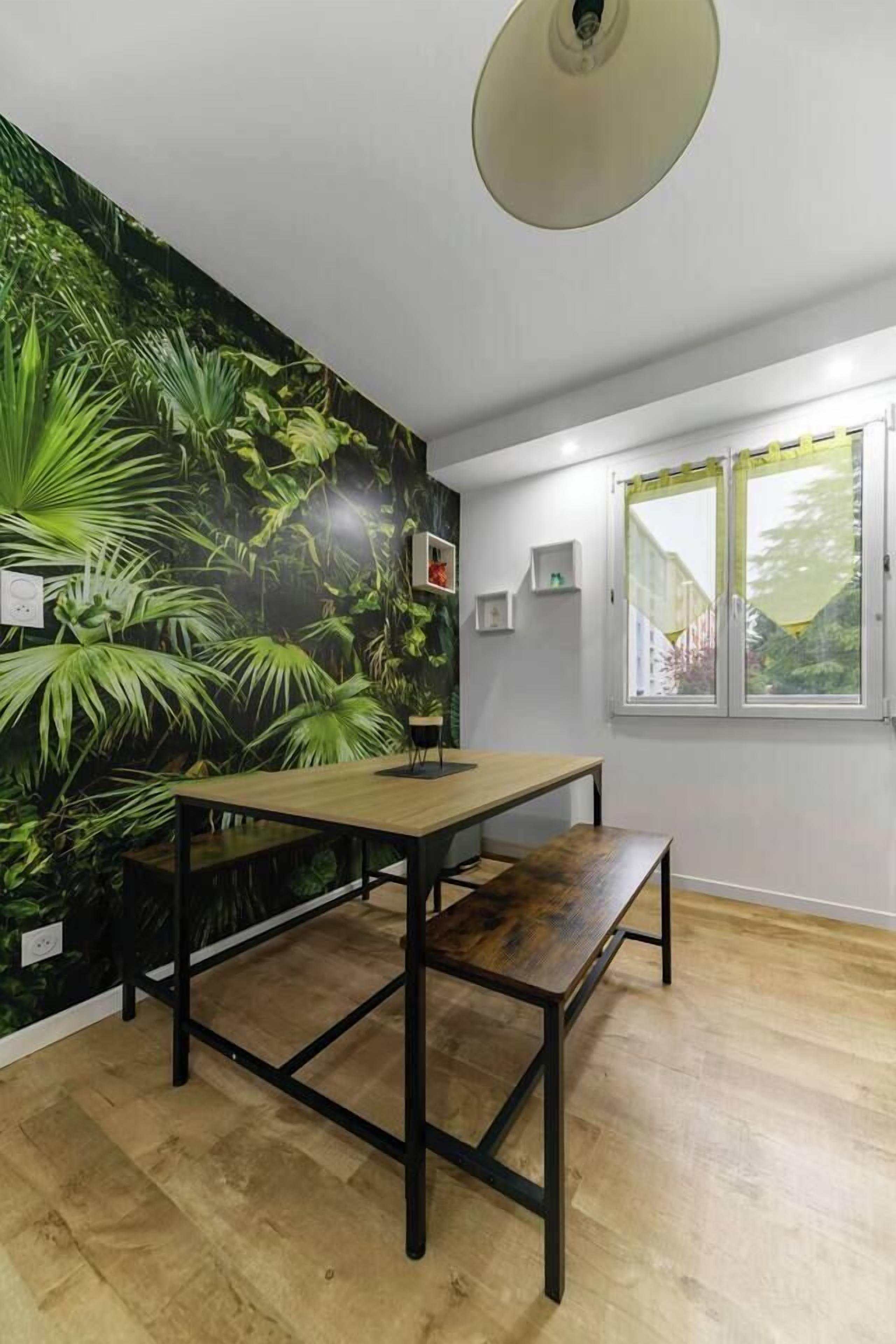 A room with a wooden floor, a table with benches, a wall mural of green foliage, a window with a view of greenery, and a ceiling light fixture.