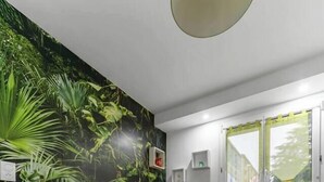 A room with a wooden floor, a table with benches, a wall mural of green foliage, a window with a view of greenery, and a ceiling light fixture. - Le Réservoir - Quiet, residential apartment (Vichy)