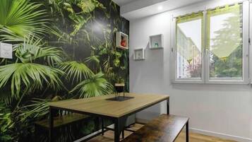 A room with a wooden floor, a table with benches, a wall mural of green foliage, a window with a view of greenery, and a ceiling light fixture.