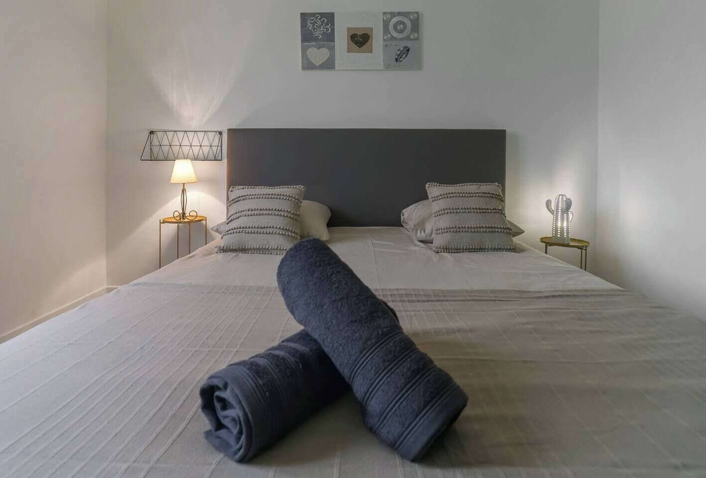 Un letto rifatto con cura, un asciugamano arrotolato, una lampada da comodino e un cactus decorativo su un tavolino.