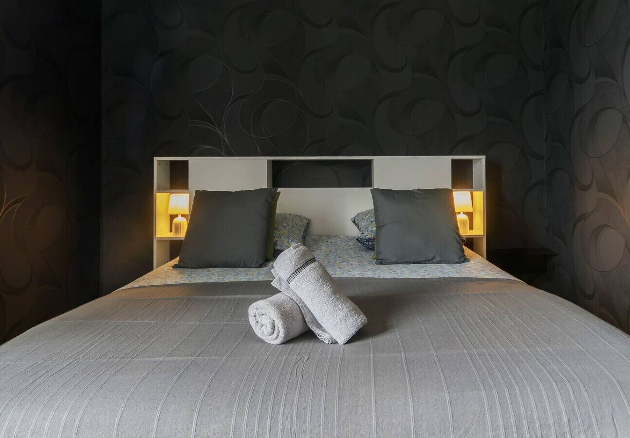 A neatly made bed with a grey bedspread and two pillows, a rolled towel, and a bedside table with a lamp.