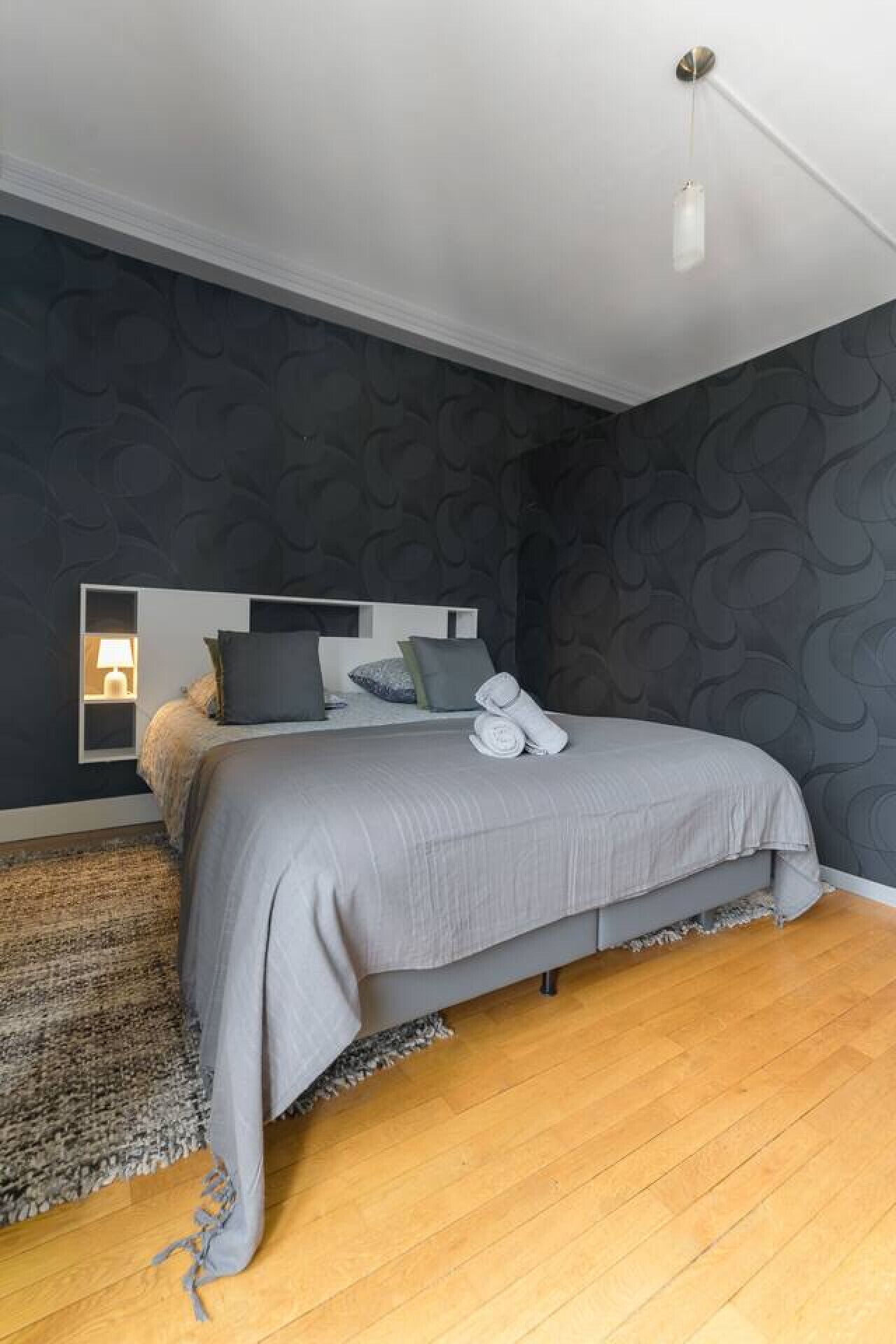 A modern bedroom with a large bed, dark patterned wallpaper, and a wooden floor.