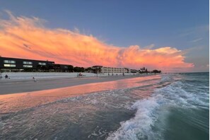 Une plage au coucher du soleil, avec des bâtiments en arrière-plan et des vagues qui se rapprochent du rivage.
