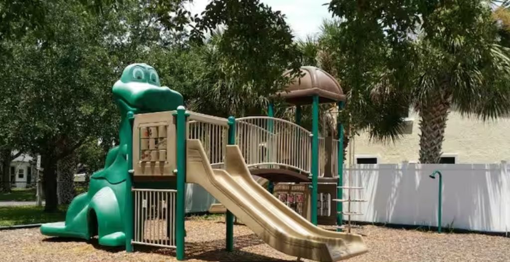A playground with a slide and climbing structures, surrounded by trees and a fence.
