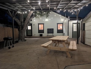 Un'area esterna coperta con tavolo da picnic, luci a stringa, un albero e un edificio con finestre.