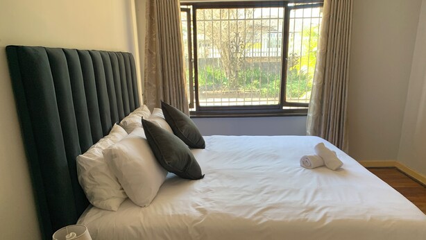 Un lit bien fait, avec du linge de lit blanc et une tête de lit vert foncé, une lampe de chevet et une vue sur la verdure à travers une fenêtre ornée de rideaux.