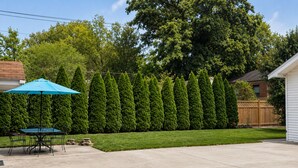 Ein Garten mit einer ordentlich geschnittenen Hecke, einem Patio-Tisch mit Stühlen und einem blauen Sonnenschirm.