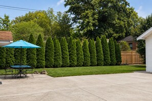 Un jardin avec une haie bien taillée, une table de patio entourée de chaises et un parasol bleu.