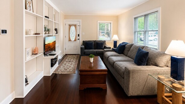 Ein Wohnzimmer mit einer Couch, Couchtisch und Fernseher auf einem Schrank. Es gibt ein Bücherregal, eine Tür mit Glasfenster und Fenster mit Jalousien.