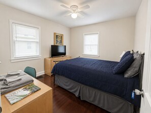 A bedroom with a bed, a desk with a television, a ceiling fan, and two windows with blinds. - Spacious Brick Home - Sleeps 8 - Near Downtown (Milford)