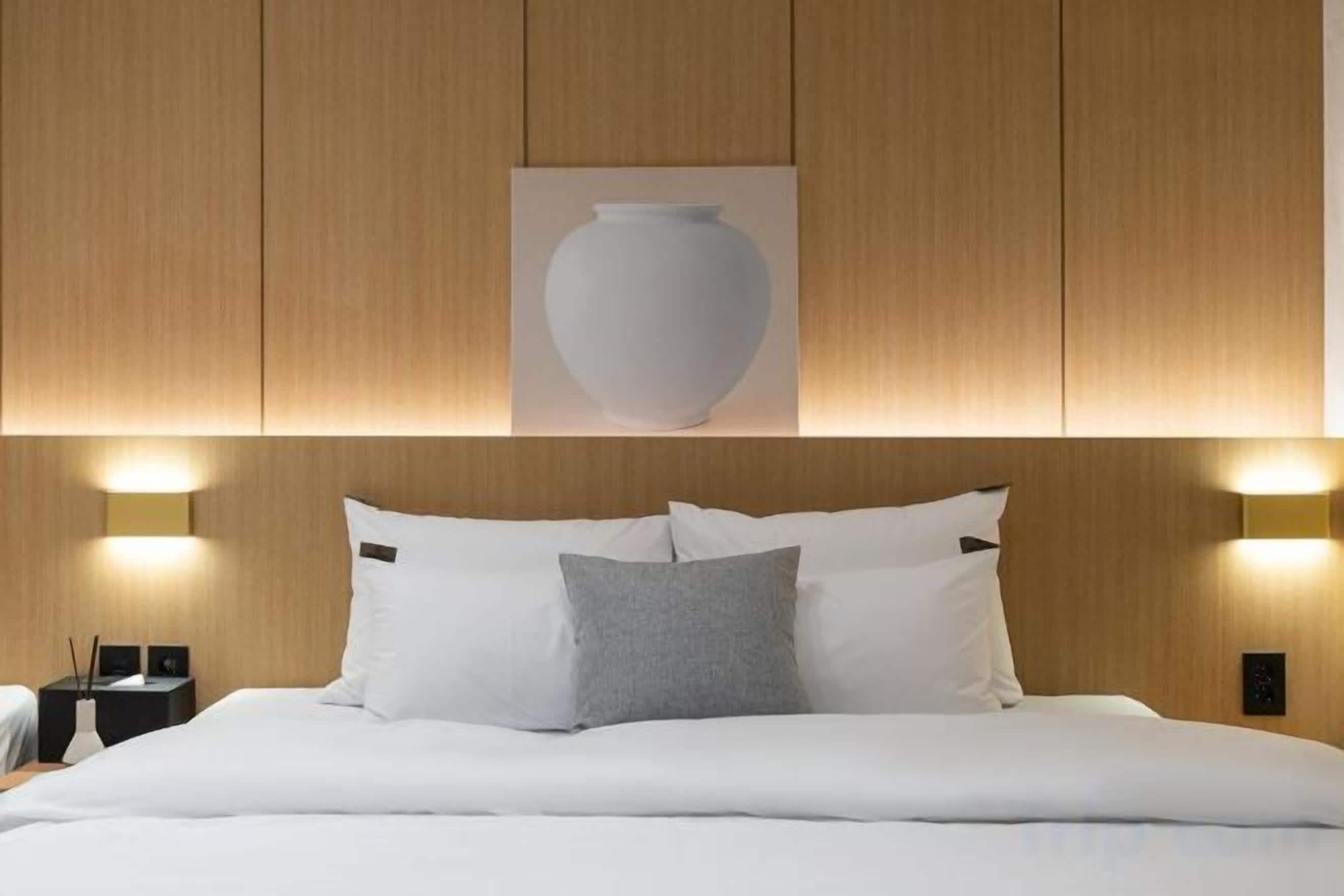 A hotel room with a bed, pillows, a wall-mounted light fixture, and a framed picture above the bed.