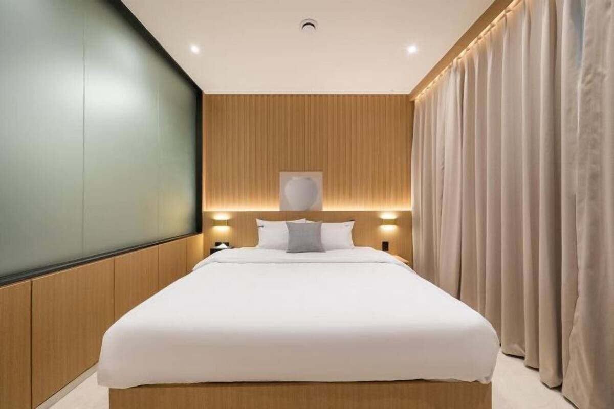 Habitación de hotel moderna con una cama grande, paneles de madera en las paredes, una lámpara en el techo y una cortina a la derecha.