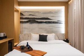 Une chambre d’hôtel avec un lit, une table de chevet en bois, un plateau contenant divers objets et une œuvre d’art accrochée au mur représentant un paysage montagneux.