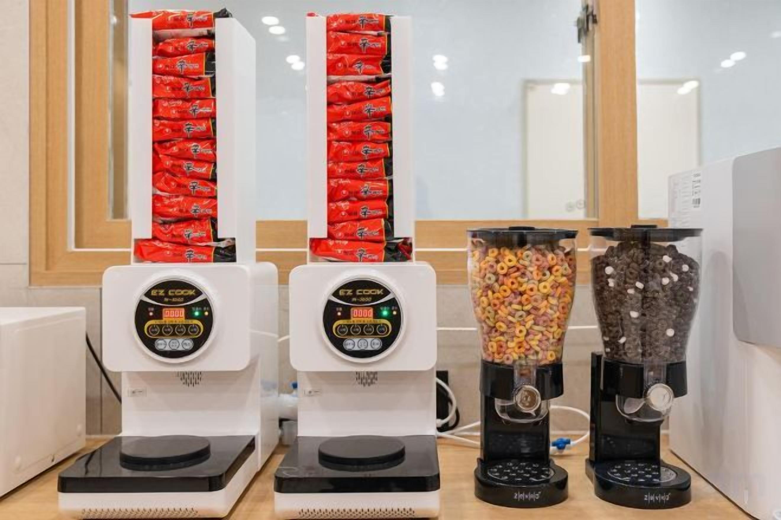 A row of four beverage dispensers with digital displays, a window with blinds, and a white wall.