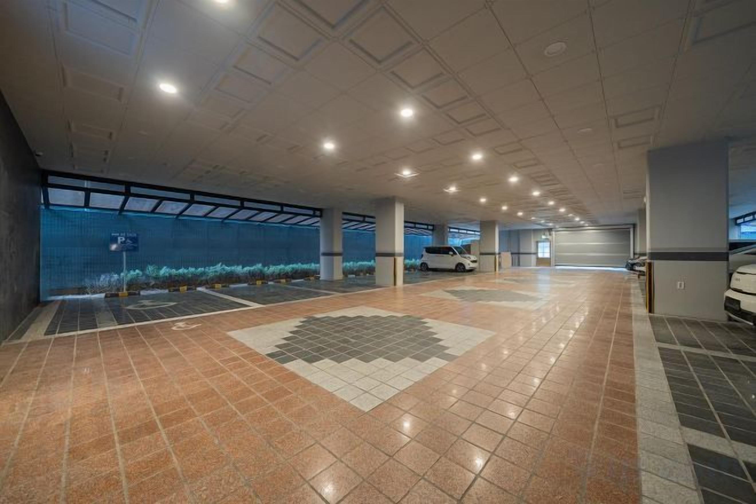 A spacious indoor parking area with a tiled floor, a white car parked, and a large window with a view of the sky.