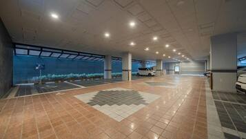 A spacious indoor parking area with a tiled floor, a white car parked, and a large window with a view of the sky.