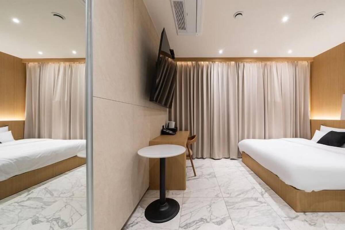 Habitación de hotel con dos camas, una mesita redonda pequeña, un televisor montado en la pared y un separador de cortina.