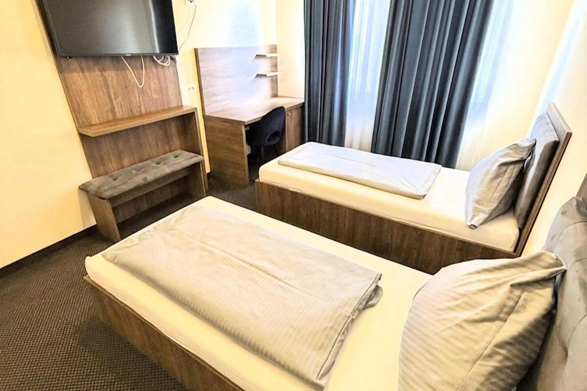 Habitació d'hotel amb dos llits, una taula de treball, una cadira, un televisor i una finestra amb cortines.