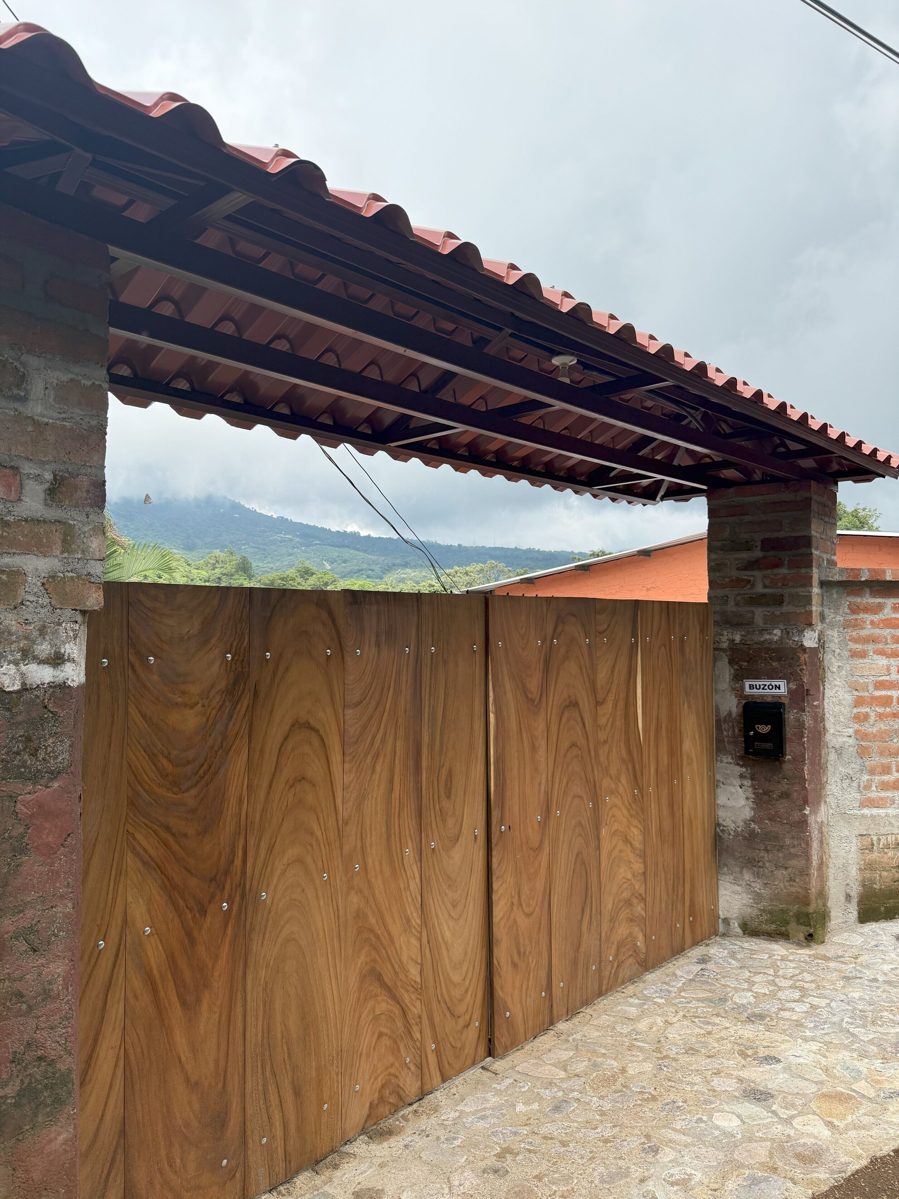 A wooden gate with a metal roof structure, a brick wall, and a stone-paved ground.