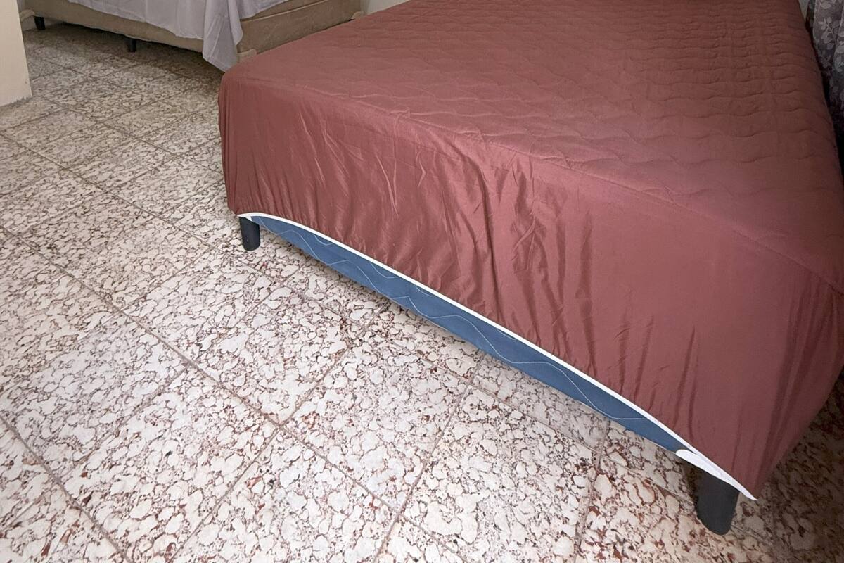 A room with two beds, one with a red bedspread and the other with a grey bedspread. The room has a tiled floor and plain walls.