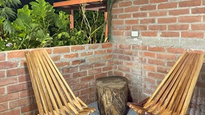 Two wooden chairs with slatted backs and metal legs, positioned on a tiled patio with a brick wall and a wooden pergola in the background. - Villa San Carlos (Juayua)